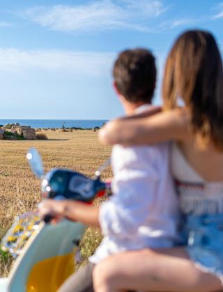 Couple on scooter admires rural landscape with hay bales and sea.